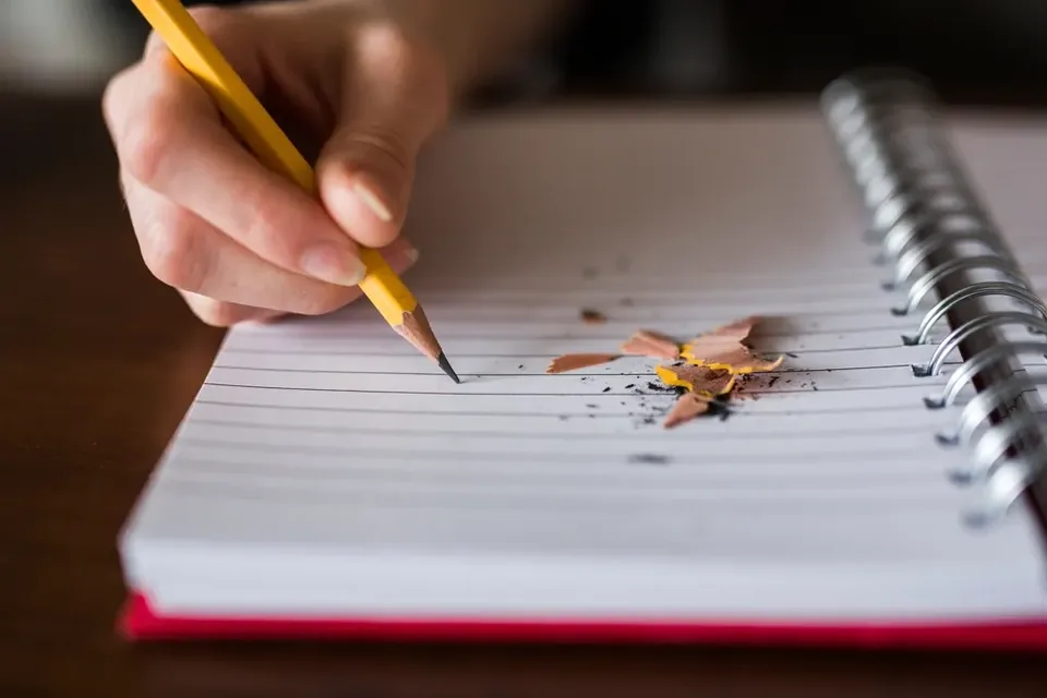 A hand writing with a freshly sharpened yellow pencil on a lined spiral notebook, shavings on the page