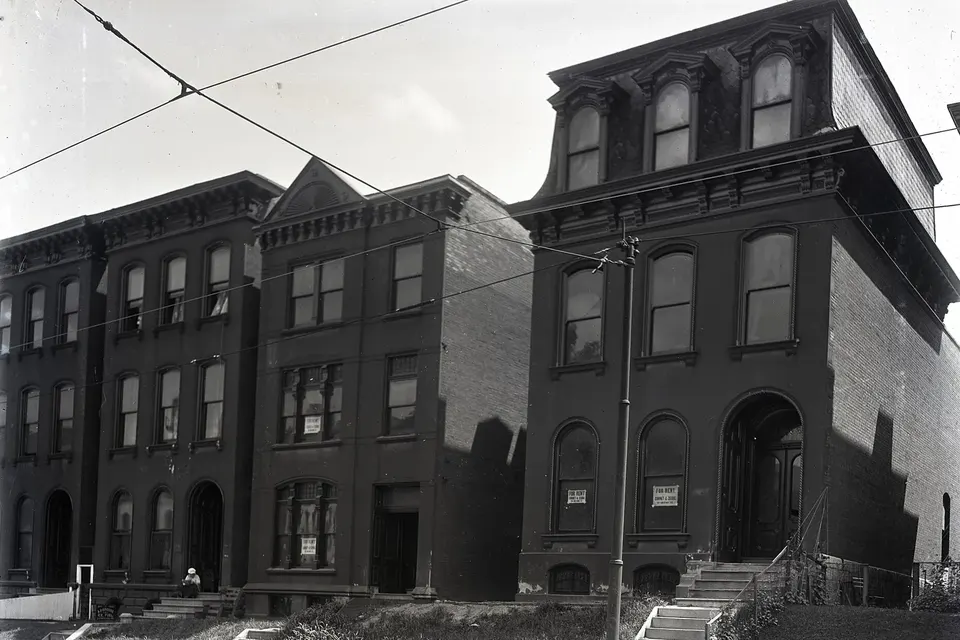 Black-and-white photograph of three-storey brick row houses with small For Rent cards in several ground-floor windows
