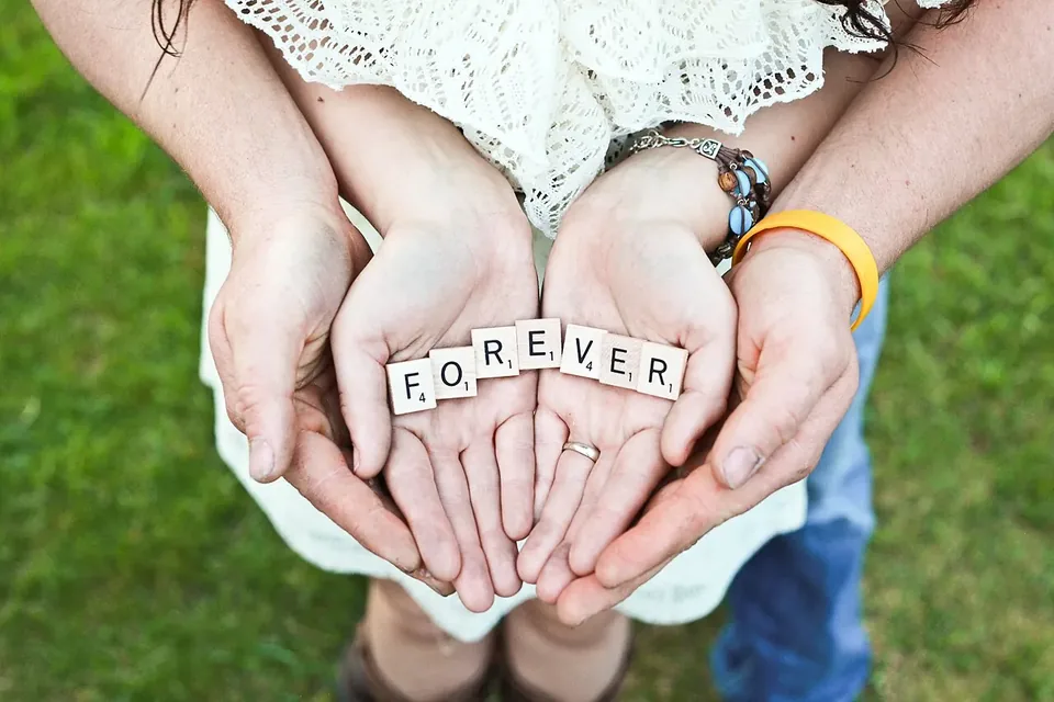 Two pairs of cupped hands holding small letter tiles that spell FOREVER, above a grassy lawn